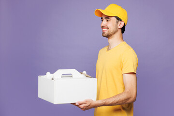 Young delivery guy employee man in yellow cap t-shirt uniform workwear work as dealer courier hold cake dessert in unmarked clear empty blank cardboard box isolated on plain pastel purple background