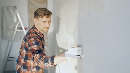 Male construction worker wearing protective gloves smoothing plaster onto wall surface during home...