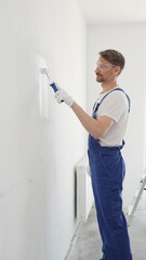 Middle aged man wearing protective white gloves and blue work overalls, applying paint with roller...