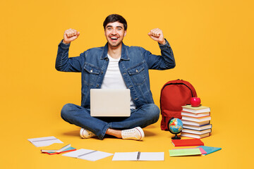 Full body young boy IT student wears jeans casual clothes sit near backpack bag books hold use work on laptop pc computer isolated on plain yellow background. High school university college concept.