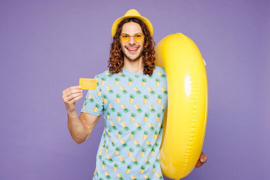 Young smiling happy man wear blue beach t-shirt yellow hat sunglasses rest near hotel pool hold rubber ring credit bank card isolated on plain purple background. Summer vacation sea sun tan concept.