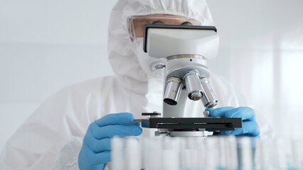 Scientific researcher wearing protective gear adjusting microscope, analyzing lab samples with...