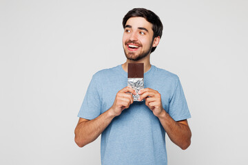 Young smiling man wear blue t-shirt casual clothes hold in hand eat chocolate bar look aside on...