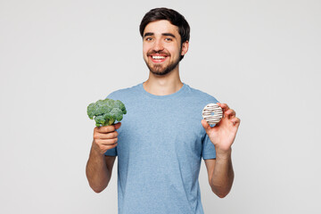 Young man wear blue t-shirt casual clothes hold in hand broccoli and donut choose between vegetable...