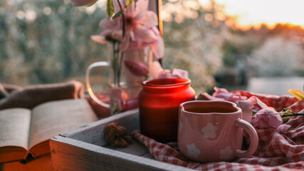 Cozy morning scene with a pink flower mug of tea, fresh flowers, an open book, and a red candle on...