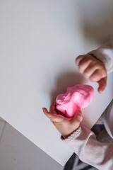 Small child squeezing soft pink modeling clay on a white table. Close-up of hands engaged in...