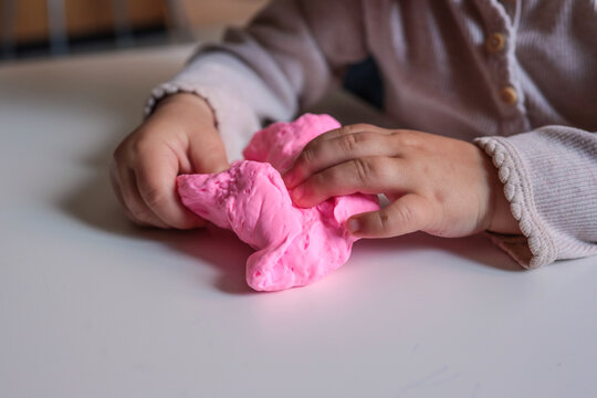 Small child squeezing soft pink modeling clay on a white table. Close-up of hands engaged in sensory and creative play, encouraging tactile exploration and motor skills