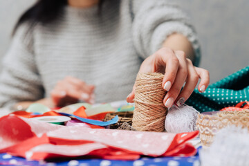 Close-up of a woman holding a roll of jute twine surrounded by colorful fabrics, ribbons, and lace on a crafting table. Creative DIY workspace with handmade decoration materials.