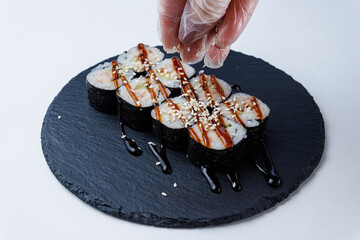 Japanese sushi rolls with seaweed and rice, drizzled with soy-based sauce and sprinkled with sesame seeds. A gloved hand adds finishing touches. Served on a black stone plate.