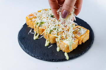 Close-up of a hand sprinkling grated cheese on deep-fried sushi rolls with green sauce on a black slate plate.