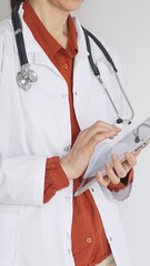 Unknown professional woman doctor, wearing white coat over a brick-red blouse and a stethoscope, is...