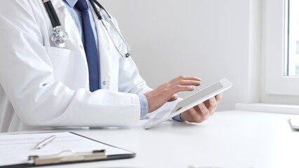 Male doctor wearing white medical coat and stethoscope is using digital tablet while sitting at...