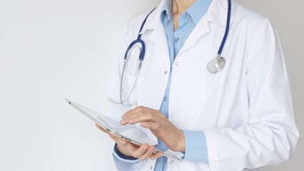 Unknown female doctor, wearing white coat over a blue blouse and a stethoscope, is using a digital...