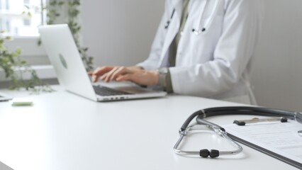 Stethoscope and medical records are on the desk opposite the doctor is typing on a laptop with a in...