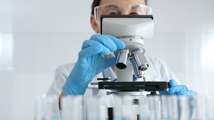 Woman science researcher wearing protective gear is checking microscope, analyzing blue test tubes...