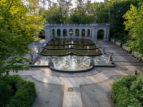 Aerial view of the M&Atilde;&curren;rchenbrunnen fountain reflecting the sky, encased by stone arches and lush green trees, Friedrichshain-Kreuzberg, Berlin, Volkspark Friedrichshain, Germany.