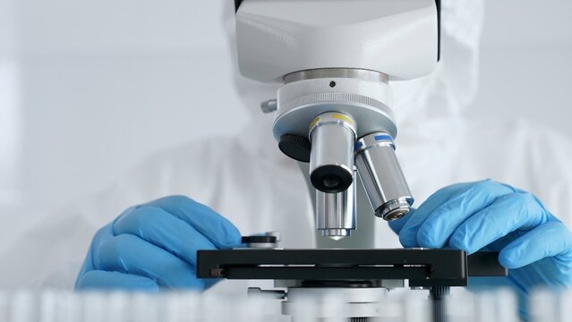Scientific researcher or virologist wearing protective gear is tuning microscope lenses opposite the lab tubes with blue liquid in laboratory setting. Medicine, healthcare and science concept