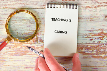 A hand holds a pencil over a notebook that displays the phrase teaching is caring near a magnifying...