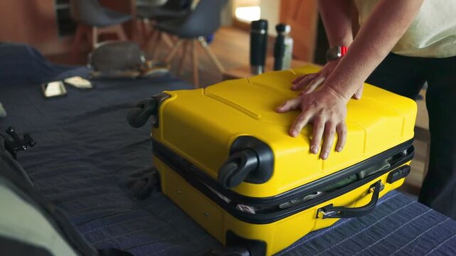 A person packs neatly folded clothes into a yellow hard-shell suitcase on a bed, preparing for a trip. The video captures an indoor travel routine, showing hands placing garments carefully