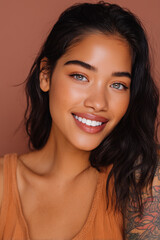 Smiling young woman with dark hair and blue eyes poses against a warm brown background, showcasing her natural beauty and stylish tattoo