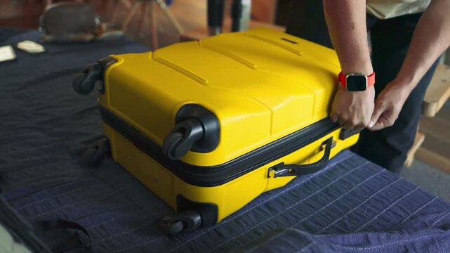 A person packs neatly folded clothes into a yellow hard-shell suitcase on a bed, preparing for a trip. The video captures an indoor travel routine, showing hands placing garments carefully