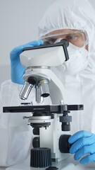Male virologist in a hazmat suit, mask, and blue gloves, diligently working at a microscope in a...