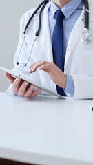 Adult male doctor wearing lab coat and stethoscope using digital tablet while sitting at desk in...