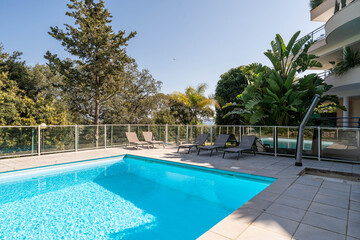 Modern residential swimming pool with tropical plants and sun loungers in sunny outdoor setting – luxury vacation lifestyle, wellness and relaxation concept