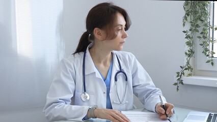 Female doctor in lab coat and stethoscope diligently writing notes on clipboard while sitting at...