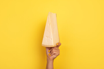  Hand with yellow nails holding a large wedge of cheese raised against a vibrant yellow background,...