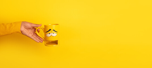  Hand in yellow sweater holding a yellow mug with a sleepy cartoon face design, isolated on a solid...