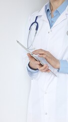 Unknown female doctor, wearing white coat over a blue blouse and a stethoscope, is using a digital...