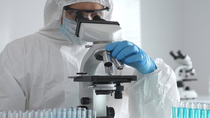 Medical virologist in protective hazmat gear examining viral samples under a microscope while...