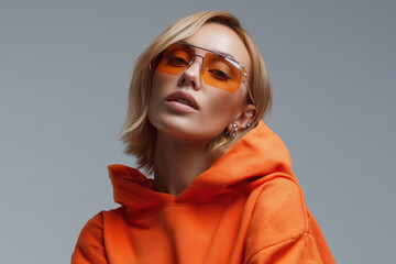 Stylish young woman in orange hoodie and trendy orange sunglasses posing confidently against gray background