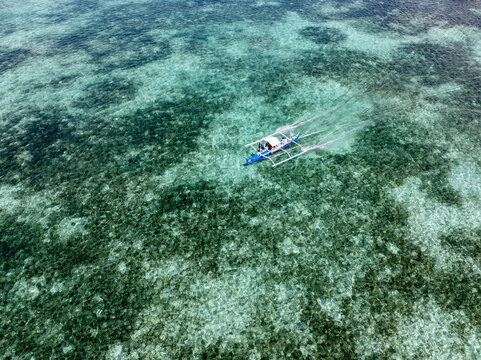 Aerial view of a traditional outrigger boat slicing through the turquoise waters, leaving a foamy wake across the vibrant coral reefs, Siargao Island, Caraga, Philippines.