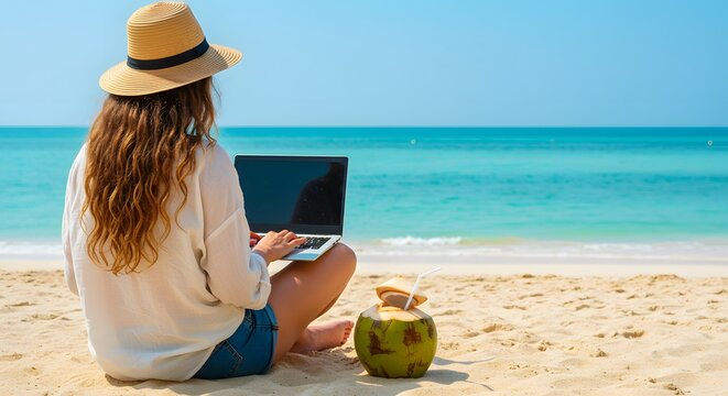 A woman wearing a straw hat works on her laptop on a sandy beach with a coconut drink beside her, enjoying the ocean view