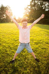 A cheerful boy jumps excitedly on a green field illuminated by the golden hour sunlight. The...