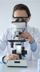 Female scientist wearing lab coat and protective glasses using microscope, analyzing samples in...