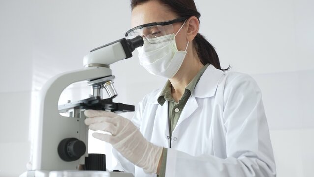 Female scientist wearing a lab coat, face mask, and safety glasses carefully adjusts a microscope, conducting research in a brightly lit laboratory setting. Medicine, healthcare and science concept