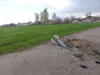 Rocket lodged in a damaged asphalt road in a countryside setting. The surrounding area shows cracked pavement, debris and green grass, with houses in the distance.
