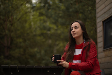 Young woman in red jacket sits outdoors with a black mug, enjoying a peaceful moment near a cabin...
