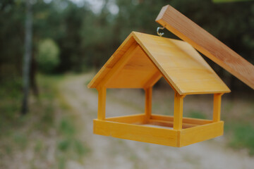Close-up of a bright yellow wooden bird feeder hanging outdoors with a forest path in the blurred...