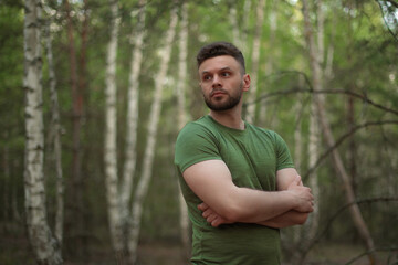 Serious young man in a green t-shirt stands in a lush birch forest with arms crossed, looking to the side. Natural lighting and soft background create a calm, confident mood.