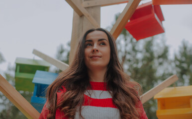 Confident young woman with long brown hair stands outdoors in front of colorful wooden birdhouses,...