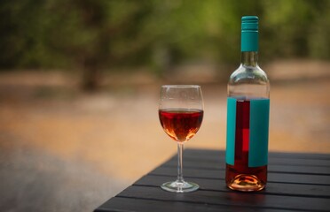 A bottle of rosé wine with a turquoise label and a filled wine glass placed on a dark wooden table outdoors. Perfect for concepts of relaxation, leisure, and outdoor dining.
