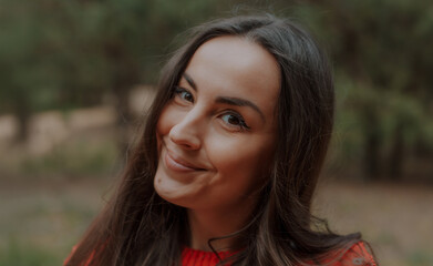 Close-up portrait of a young woman with long brown hair smiling warmly at the camera outdoors....
