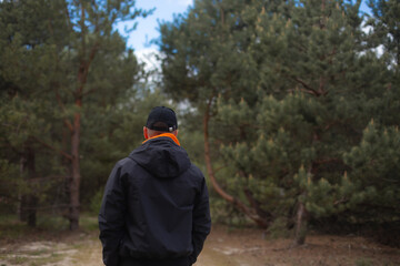 Man standing with his back to the camera on a forest path, wearing a black jacket and cap with an orange hoodie, surrounded by tall pine trees. Concept of solitude and reflection outdoors.