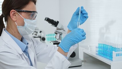 Female scientist wearing a face mask and blue gloves, carefully dropping liquid from a pipette into...