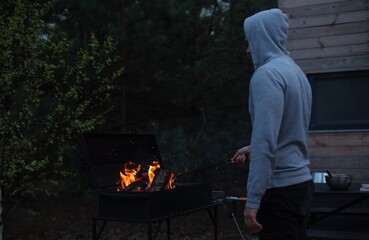 Man in a gray hoodie tends to a fire in a black grill outside a wooden cabin during dusk. Glowing...