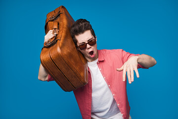 A trendy young man holding a vintage suitcase, wearing sunglasses and a bright outfit, poses with...
